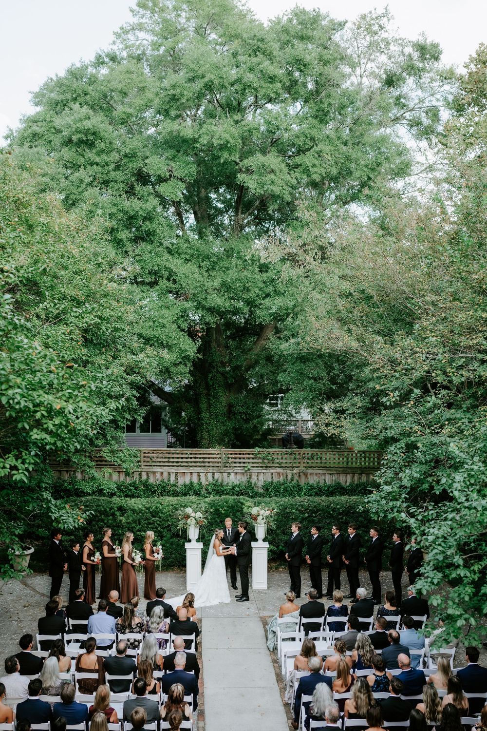 Outdoor garden wedding ceremony with bride and groom at the altar, flanked by bridesmaids and groomsmen under tall trees.