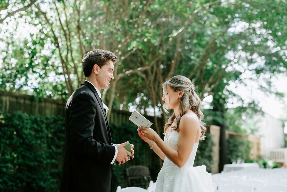 Bride reads wedding vows from paper to groom in black tuxedo during outdoor garden ceremony.