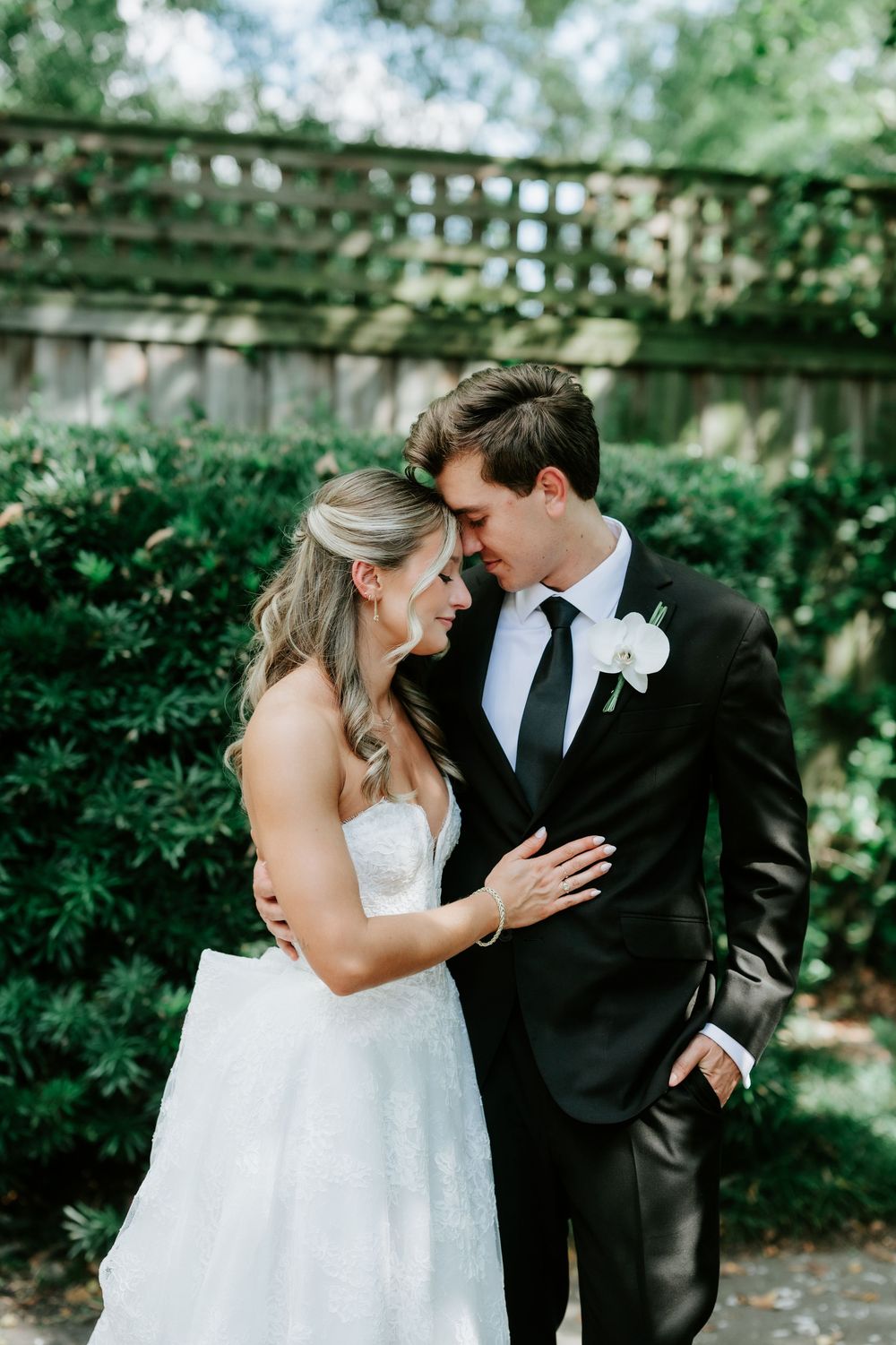 Bride and groom share an intimate forehead touch moment in a lush green garden, both dressed in elegant wedding attire.