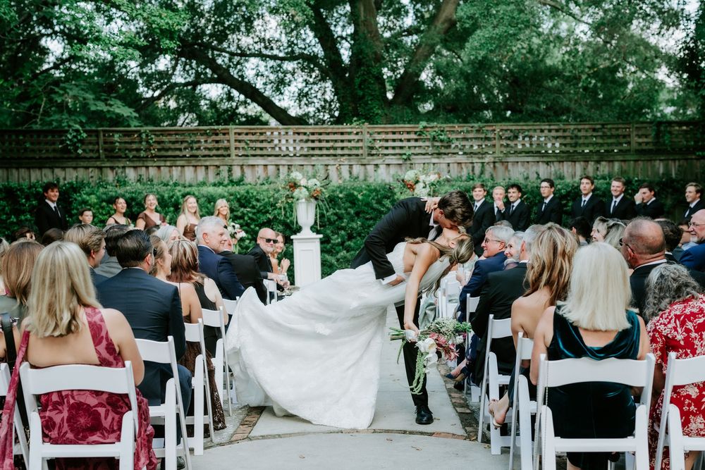 Groom dips bride at outdoor wedding ceremony altar while guests watch from white chairs in a lush garden venue.