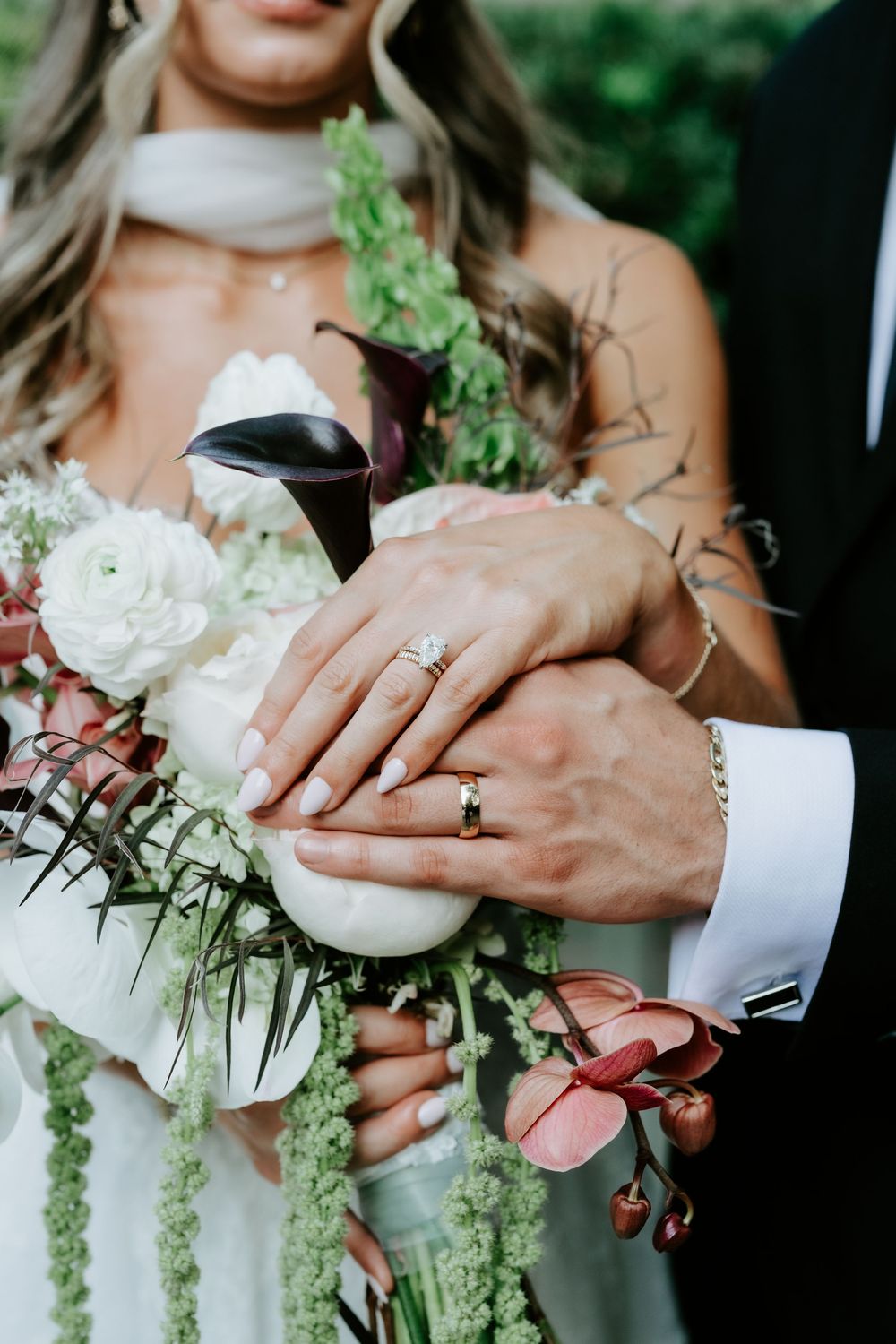 Close-up of bride and groom hands with wedding rings resting on a lush bridal bouquet of white and burgundy flowers.