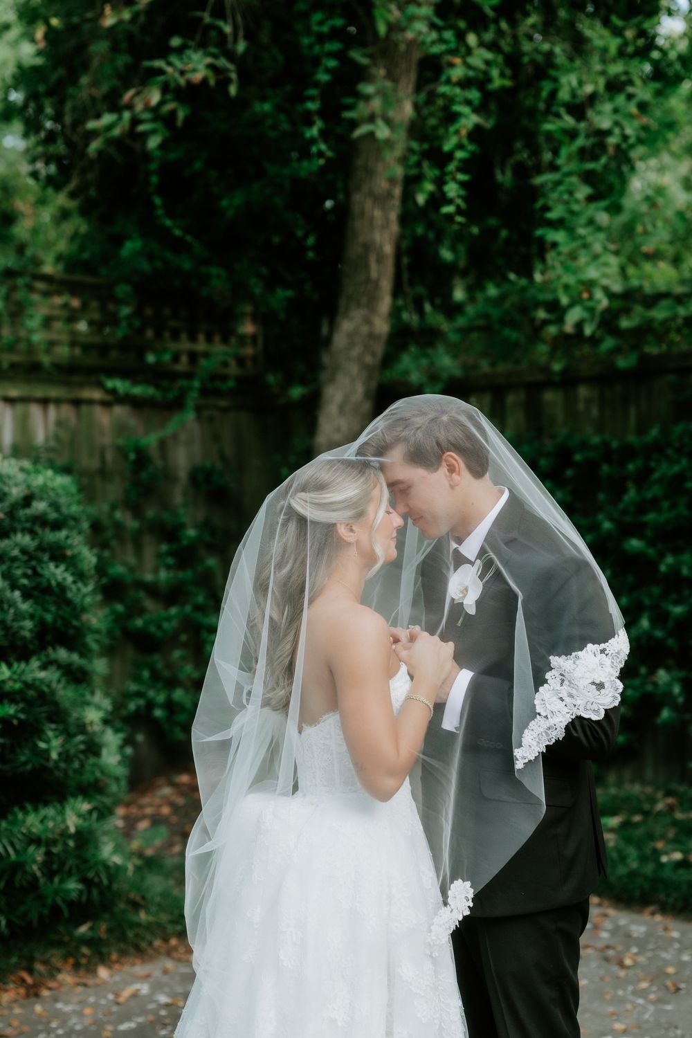 Bride and groom share intimate moment under wedding veil in lush green garden setting.