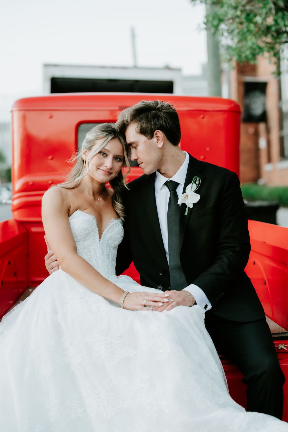 Bride and groom share a romantic moment leaning together beside a vintage red truck at their wedding.
