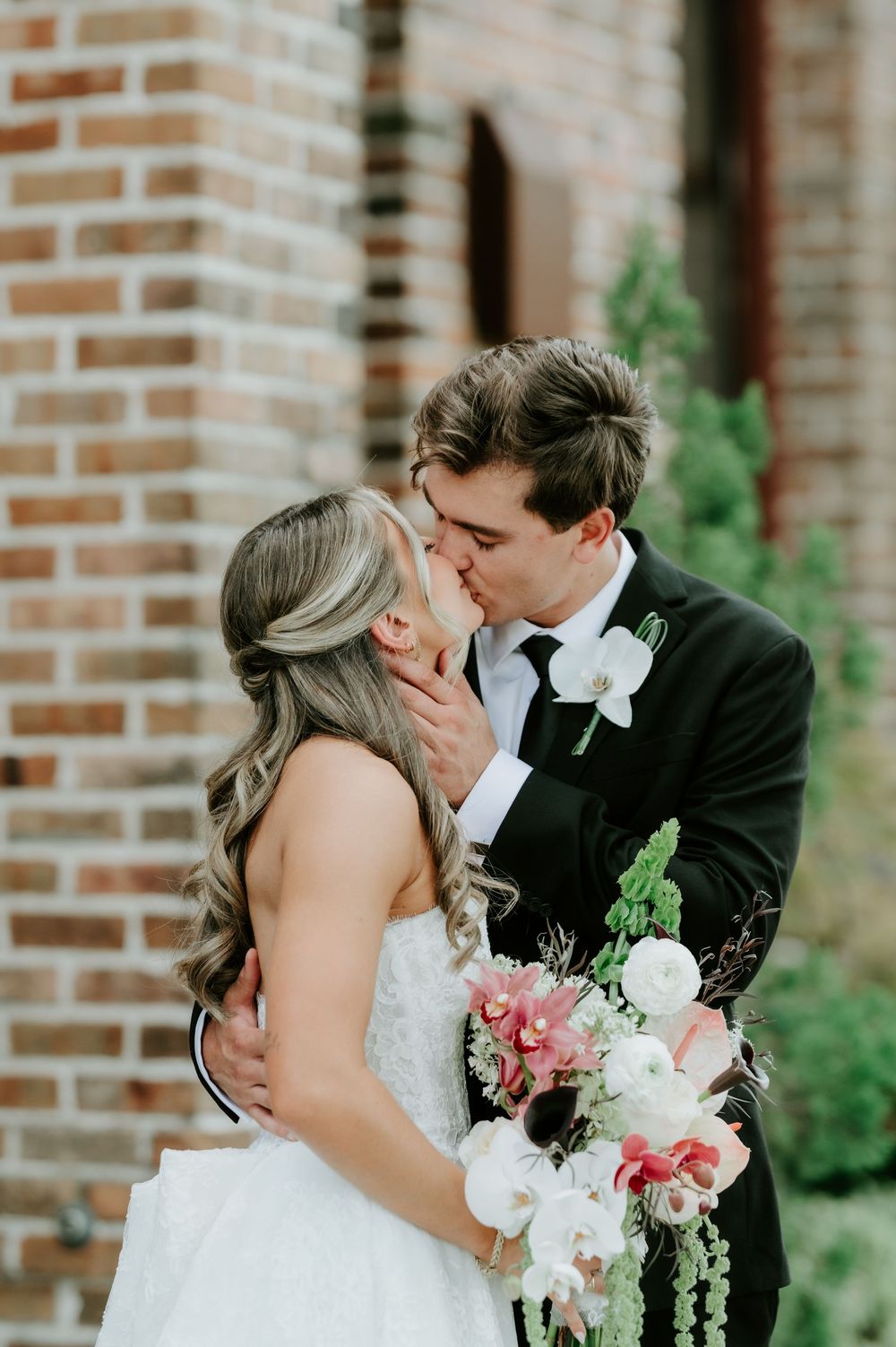 Bride and groom share a kiss near a brick wall, bride holding a cascading pink and white floral bouquet.