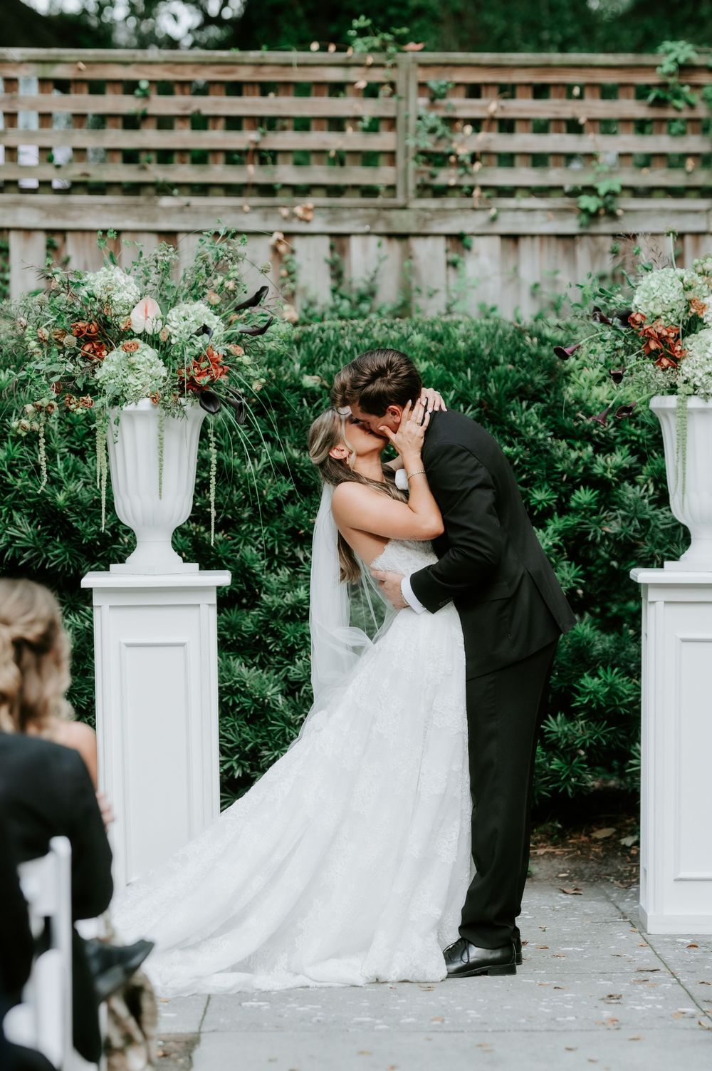 Bride and groom share first kiss at outdoor garden wedding ceremony with white pedestals and floral arrangements.