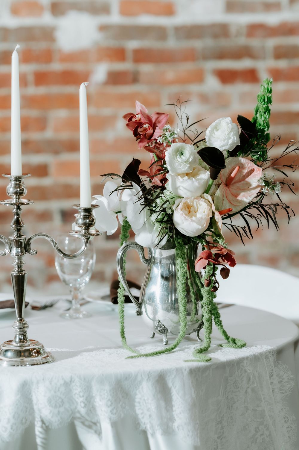 Elegant wedding table centerpiece featuring white and pink roses in a silver pitcher beside silver candelabra on lace linen.