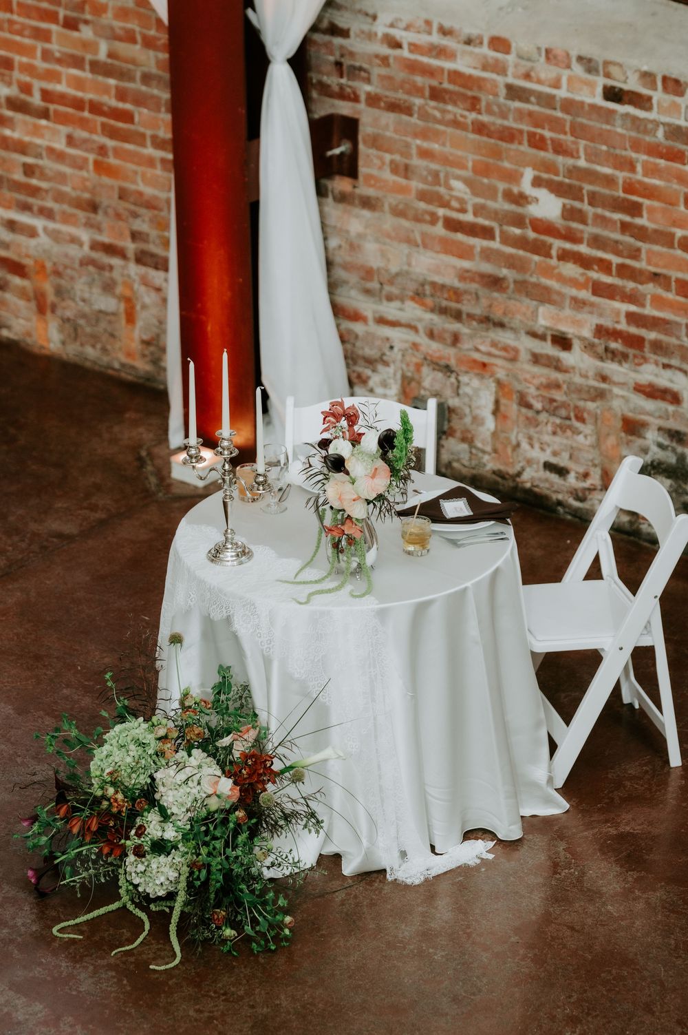 Elegant sweetheart table with white linen, taper candles, floral centerpiece, and lush bouquet on exposed brick warehouse floor.