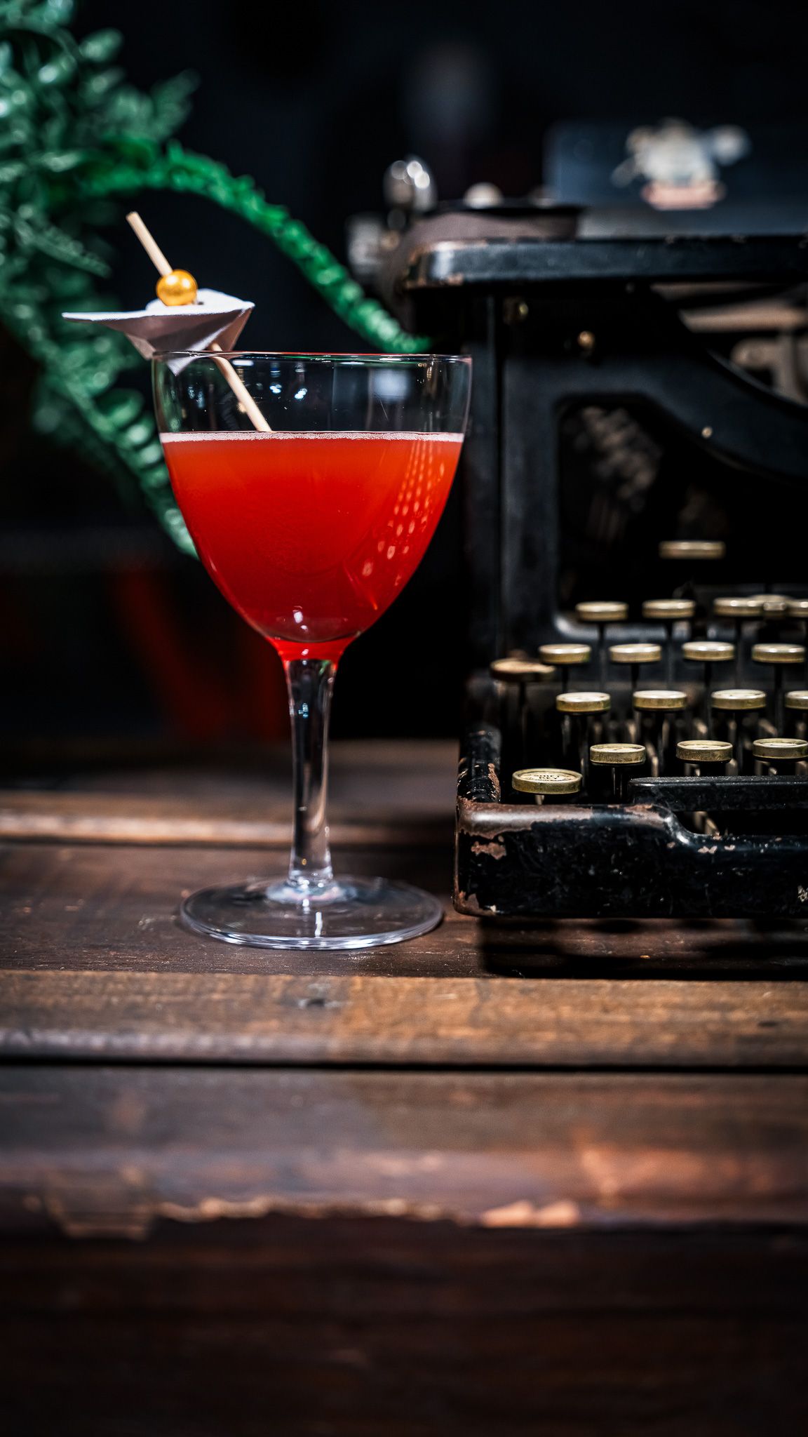 Red cocktail in a coupe glass next to vintage typewriter on rustic wooden surface with moody lighting.