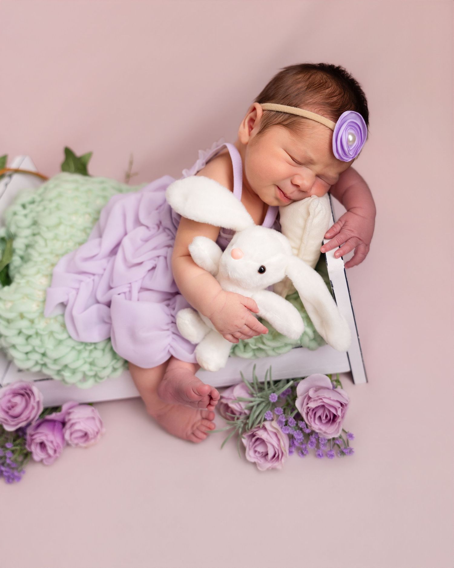 Sleeping newborn baby girl in lavender dress holding white bunny stuffed animal with purple flowers and green knit blanket.