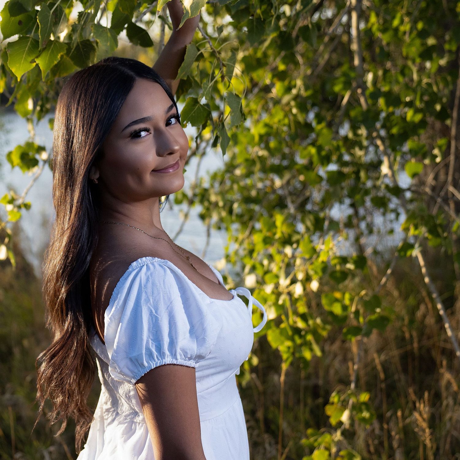 Young woman in white dress smiling outdoors among green trees in warm sunlight.