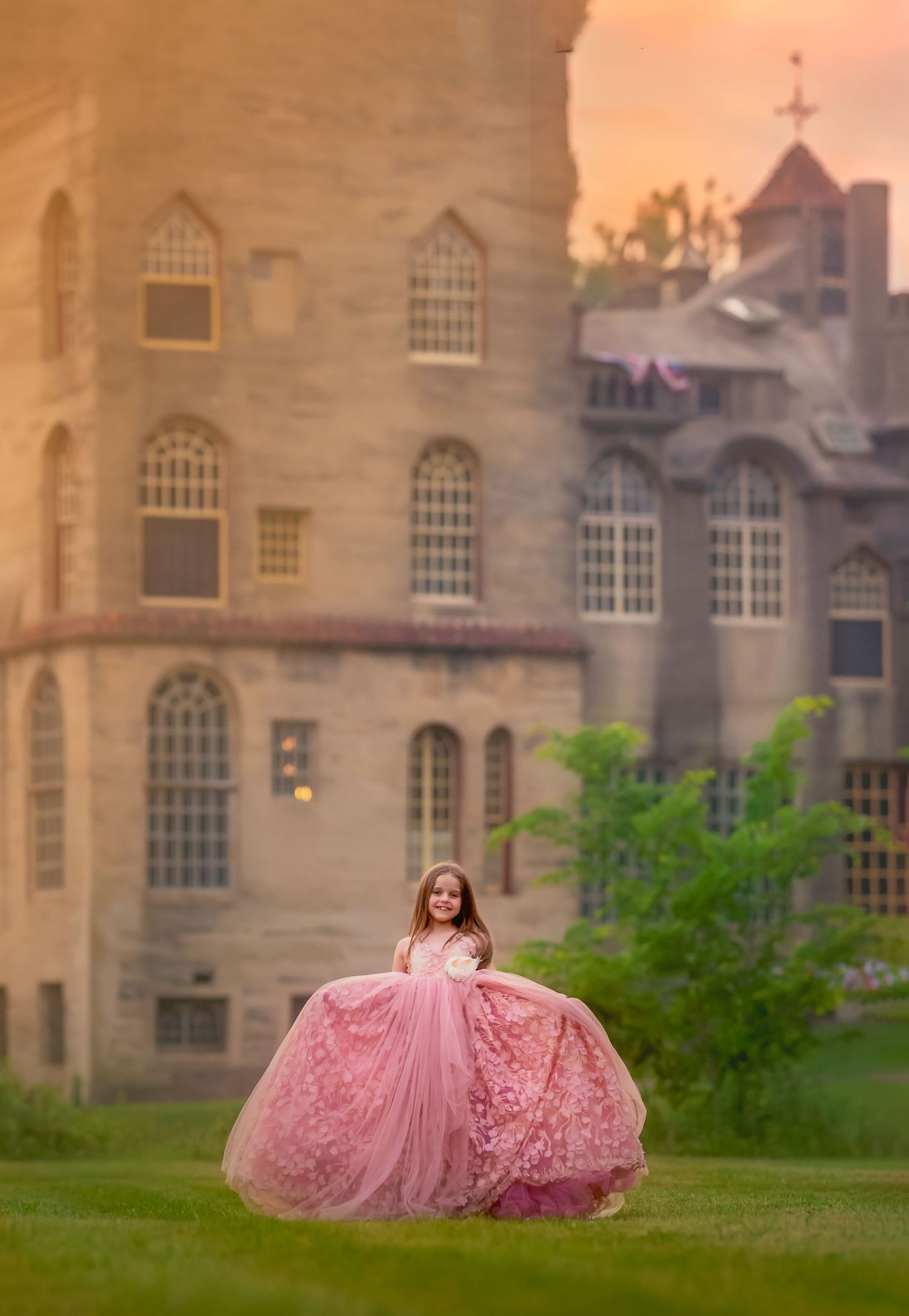 Young girl in a pink princess ballgown poses on a castle lawn at sunset.