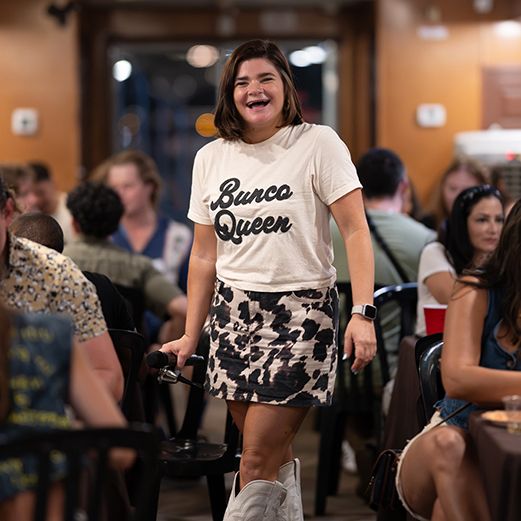 Smiling woman in a Bunco Queen t-shirt and cow print skirt walks through a lively restaurant with diners seated at tables behind her.