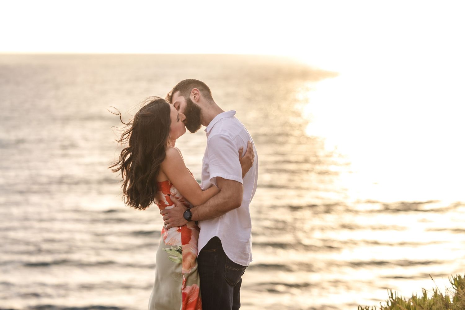 Couple embracing while standing on a coastal cliff overlooking the ocean surrounded by golden wildflowers.