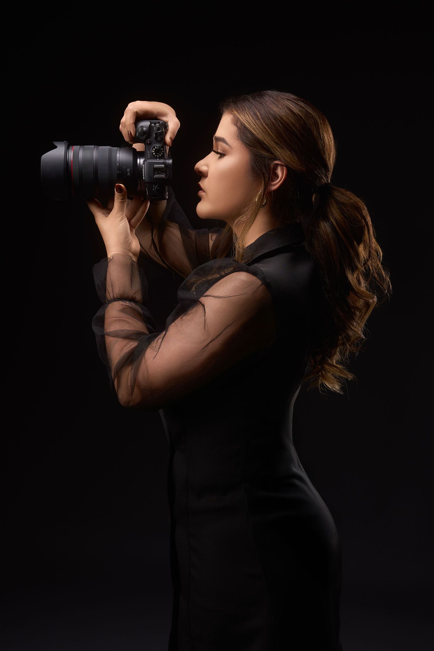 A young woman in a black mesh dress holds a DSLR camera up to her eye, photographing against a dark background.