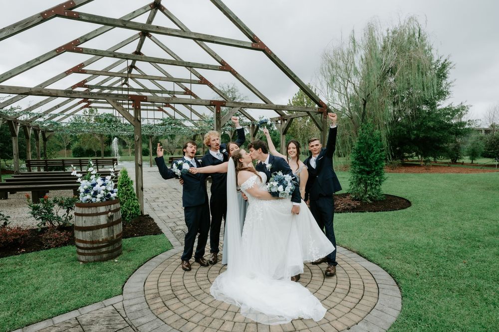 Bride in a white gown kisses her groom under a wooden pergola while groomsmen cheer in a garden wedding ceremony.