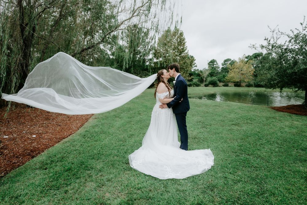 A bride and groom kiss on a green lawn near a pond as her long sheer veil flows dramatically in the wind.
