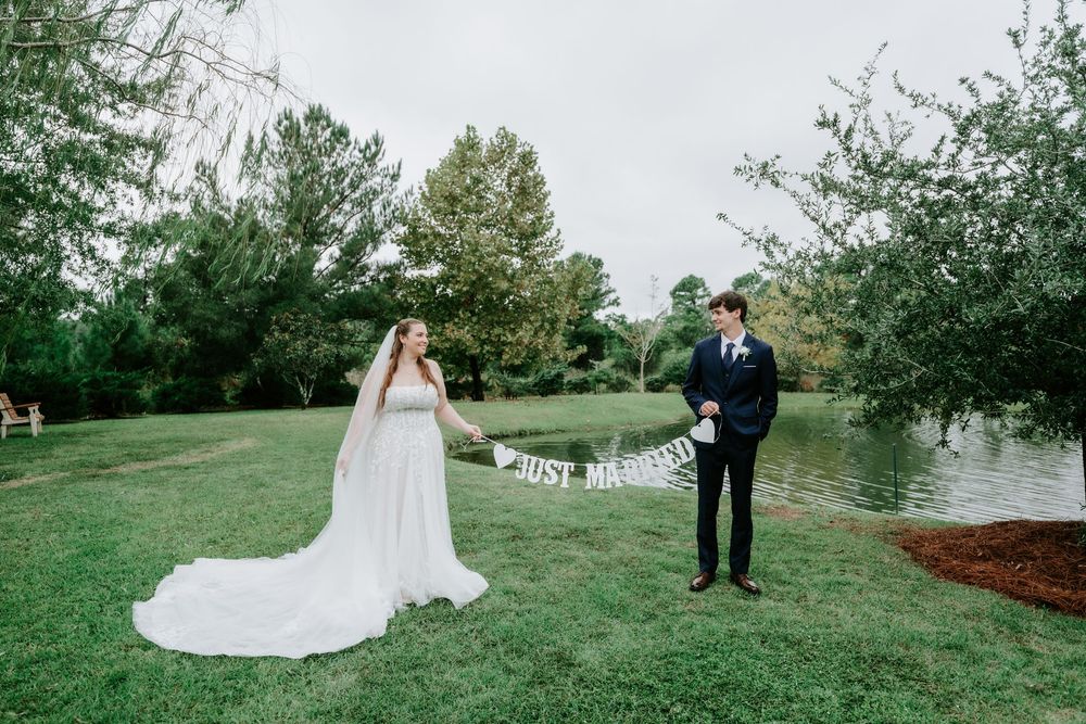 Bride in a long white gown and veil and groom in a dark suit hold a just married banner on a green lawn by a pond.