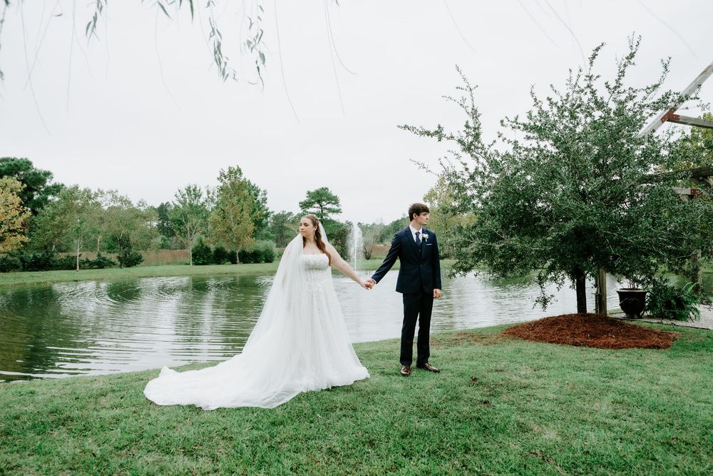 Bride in a flowing white gown holds hands with a groom in a dark suit on a grassy lawn beside a tranquil pond and willow trees.