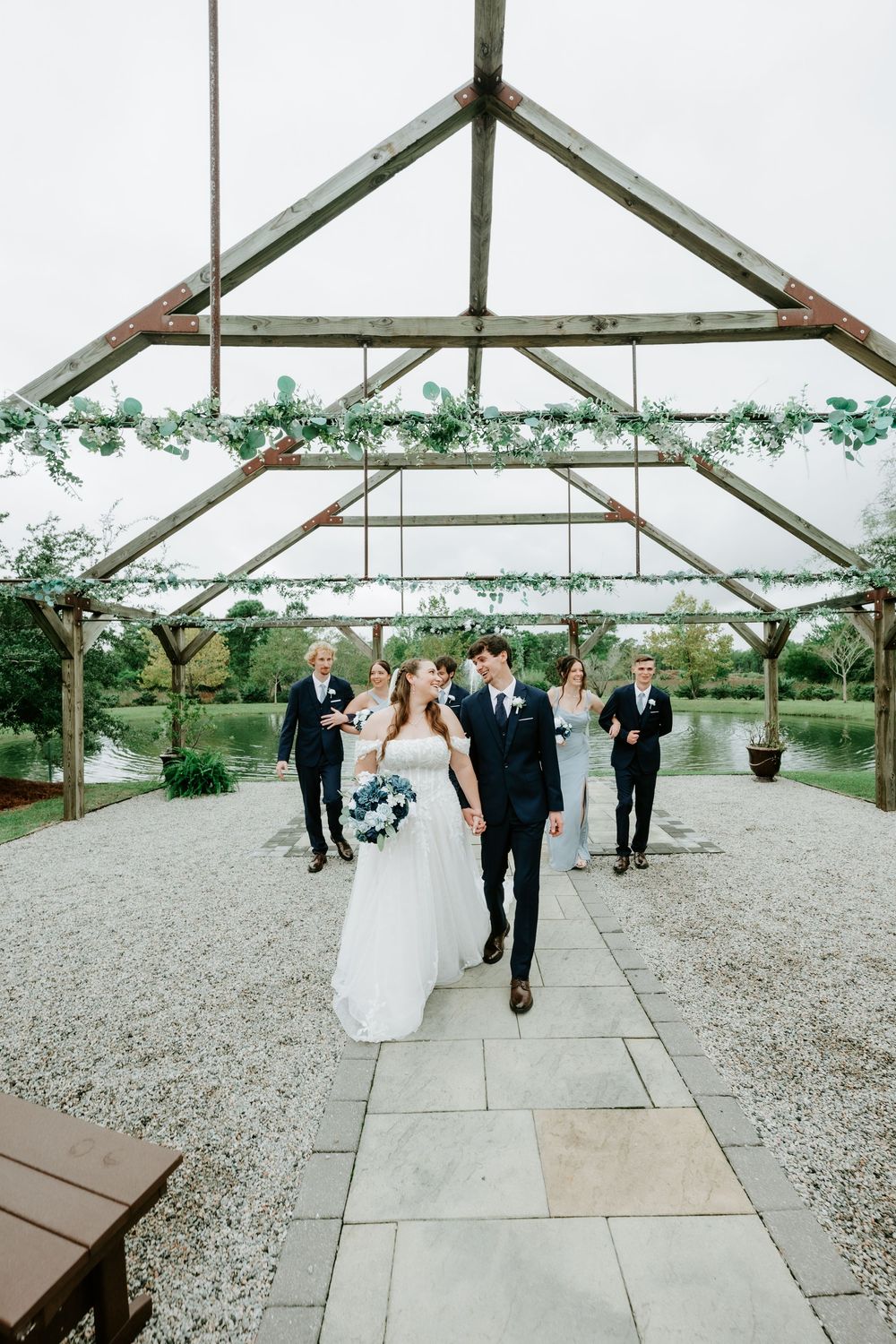 Bride and groom walk hand in hand down a stone path beneath a rustic wooden pergola draped with greenery, followed by their wedding party.