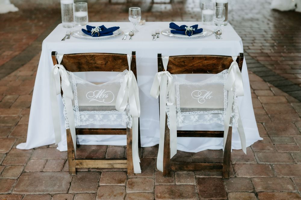 Wedding sweetheart table with two wooden folding chairs draped in white lace covers, navy napkins and glassware on a white linen tablecloth.