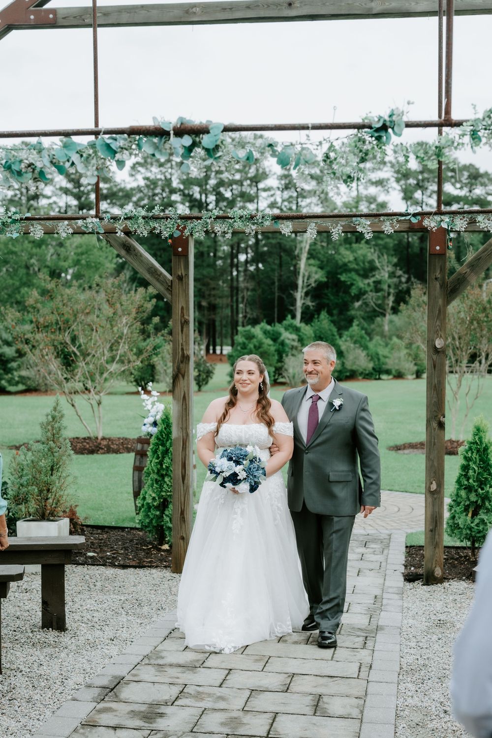 A father in a gray suit walks his smiling bride in a strapless gown down a stone path toward a rustic outdoor wedding arbor.