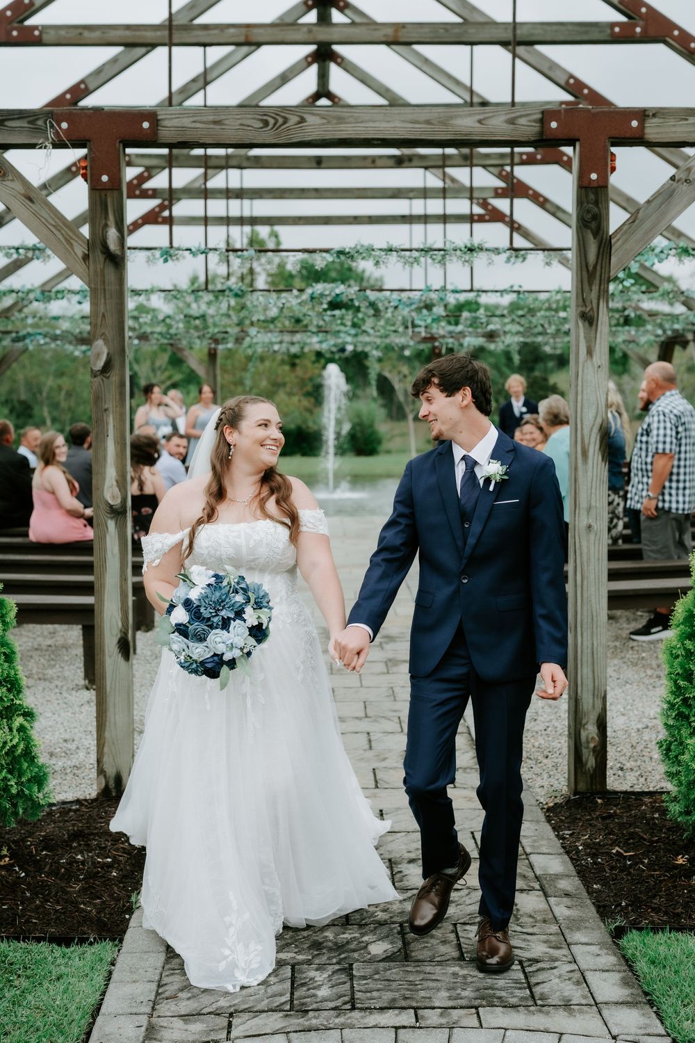 Smiling bride in a white lace gown holding a blue bouquet walks hand in hand with her groom in a navy suit under a wooden pergola.