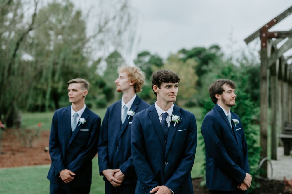 Four groomsmen in navy suits with boutonnieres stand in a row outdoors at a garden wedding, with trees and a wooden pergola behind them.