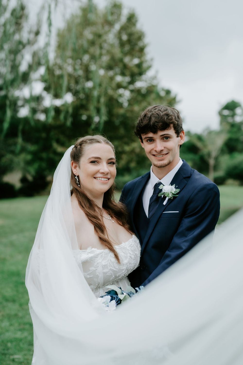 Smiling bride in a lace wedding gown and veil poses beside her groom in a navy suit with a white boutonniere on a green lawn.