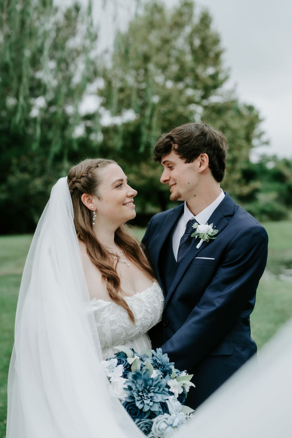 A bride in a lace gown and veil smiles at her groom in a navy suit while holding a blue and white floral bouquet outdoors.