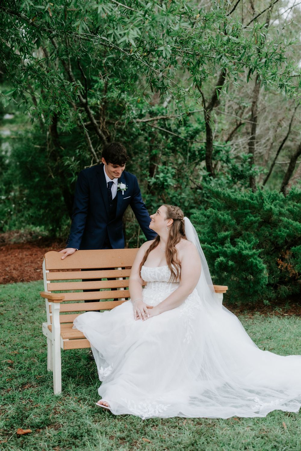 Bride in a white lace gown and veil sits on a wooden bench smiling up at the groom in a navy suit, surrounded by greenery.