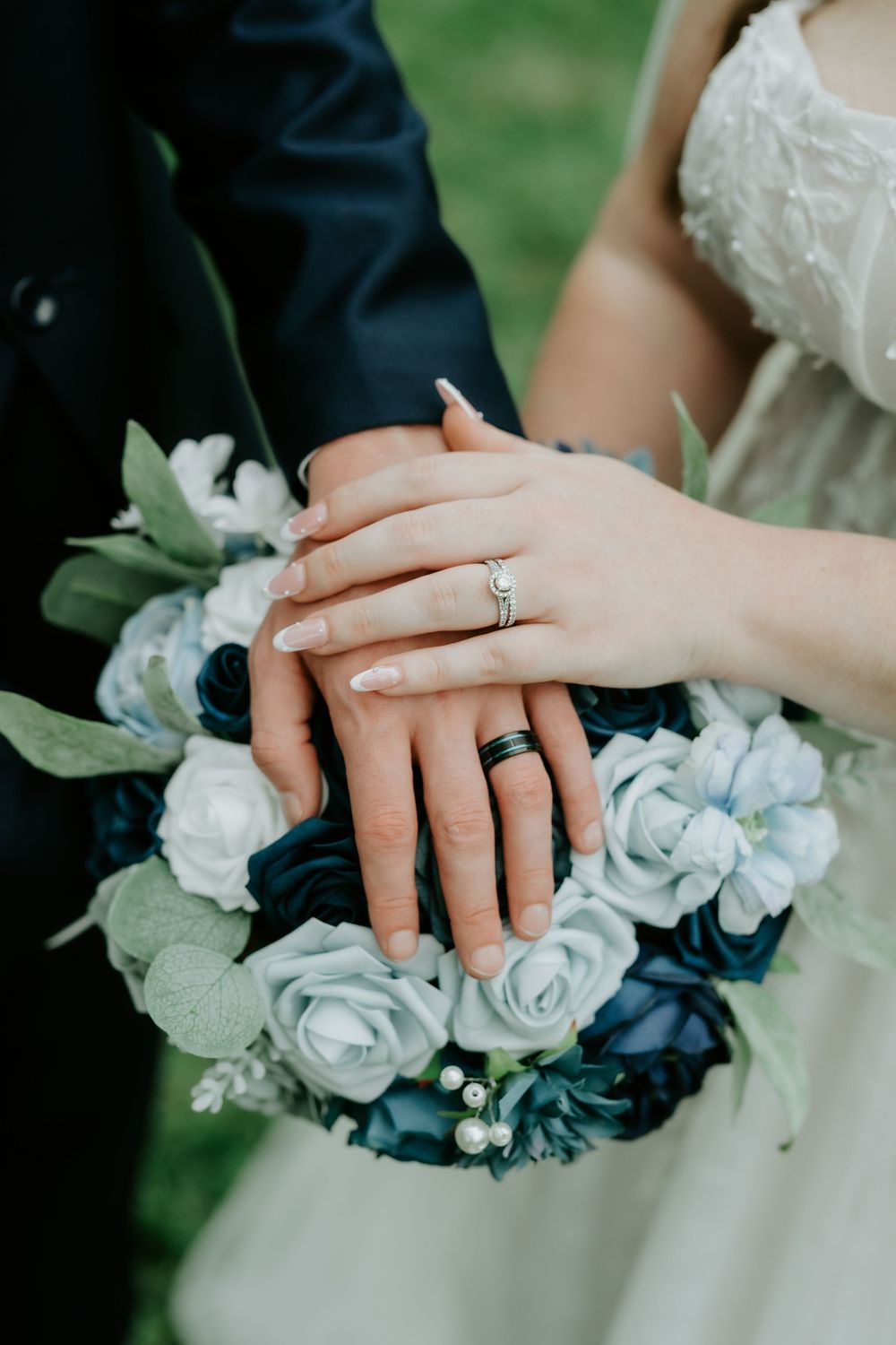 Close-up of a bride and groom's stacked hands showing wedding rings over a bouquet of white and navy blue roses with greenery.