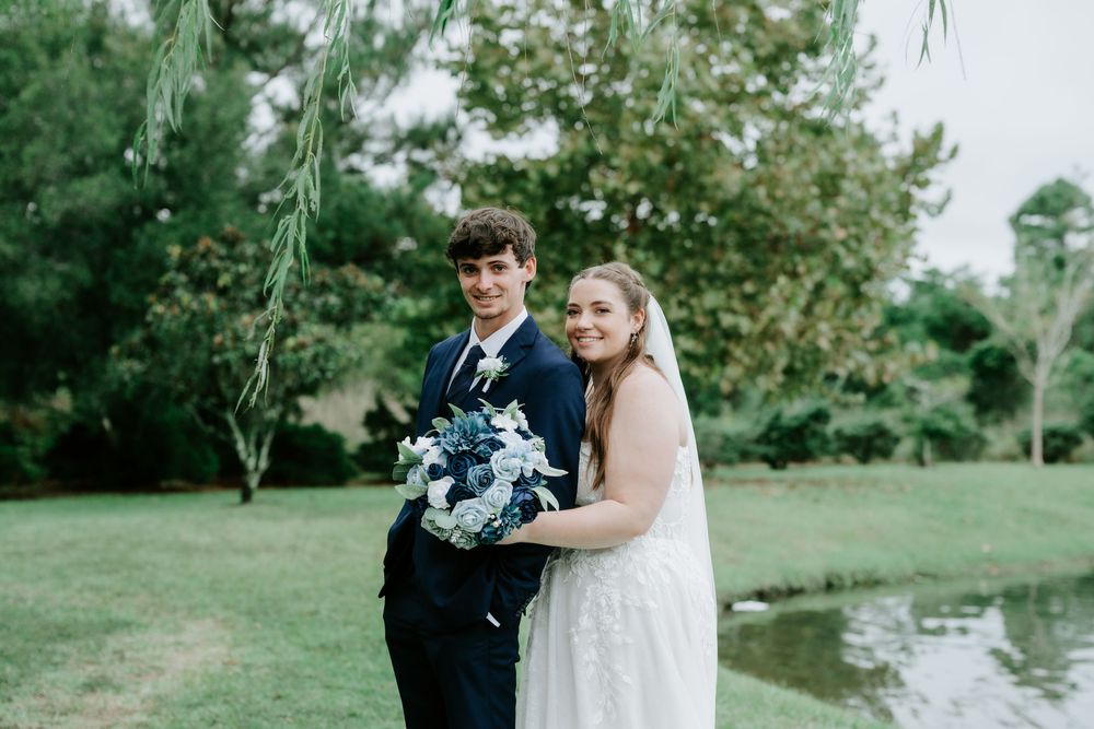 Smiling bride in a white gown and veil holds a blue and white bouquet beside her groom in a navy suit on a sunny, tree-lined lawn.