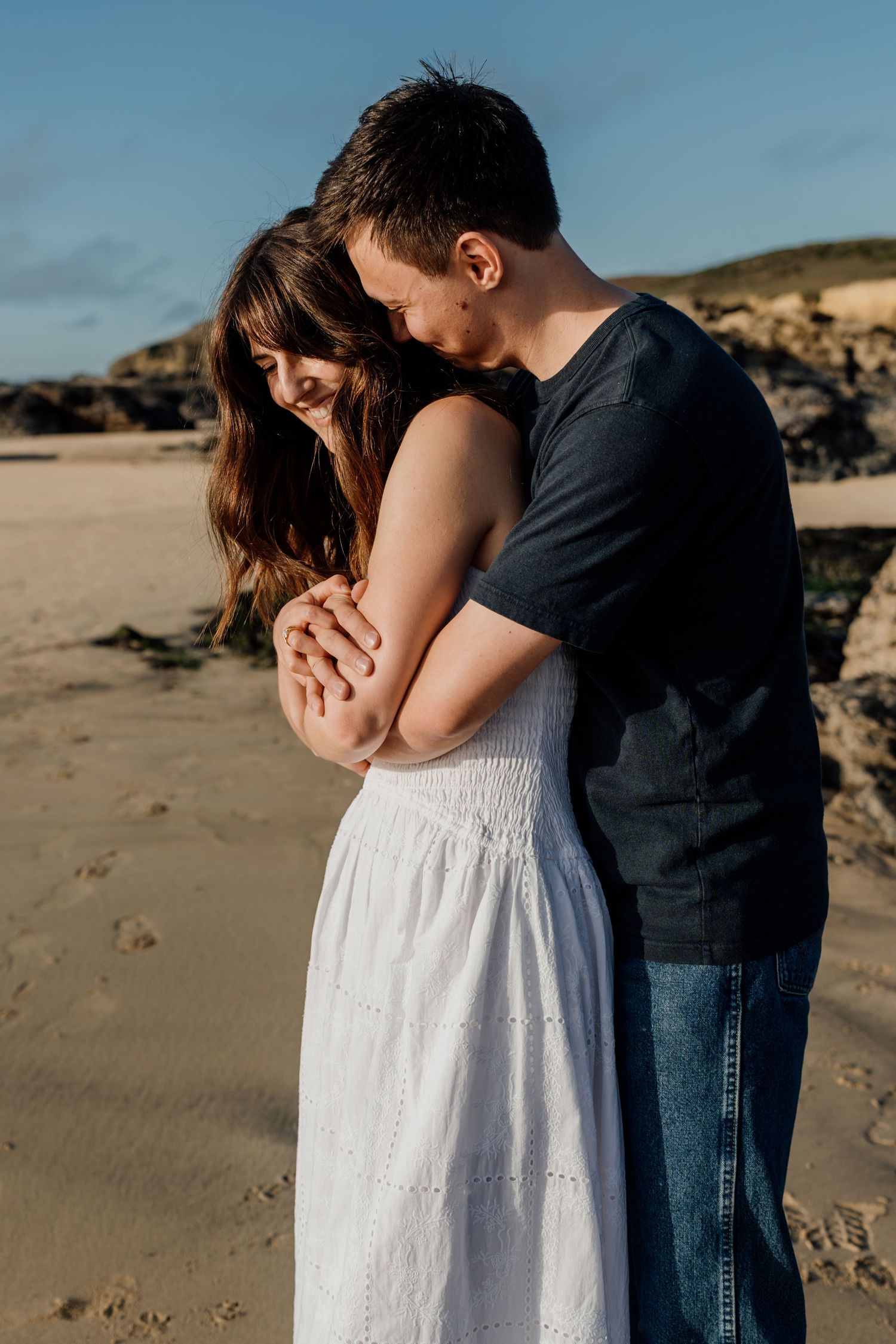 Couple holding hands on a reflective beach at golden sunset, woman in white dress, waves in background.