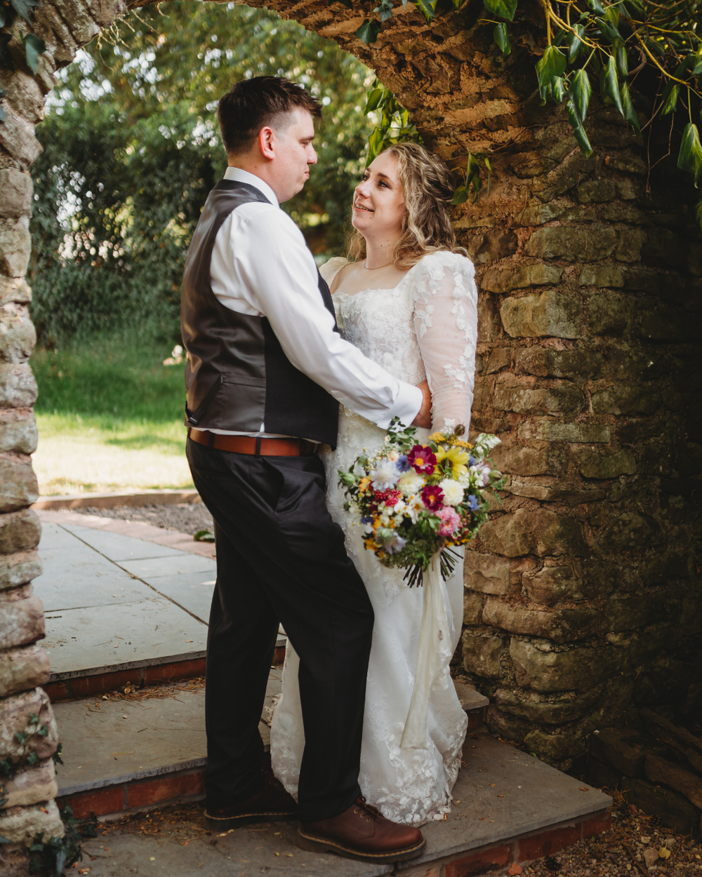 Bride and groom share a romantic moment under a stone arch, bride holding a colorful wildflower bouquet in a lace wedding dress.