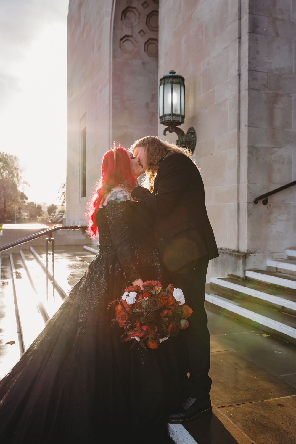 Gothic bride in black dress with red hair kisses groom outside stone building, holding dark floral bouquet.