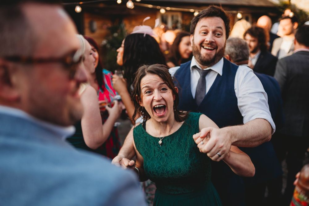 Joyful wedding guests dancing and laughing at an outdoor evening reception, woman in green dress and man in blue vest.