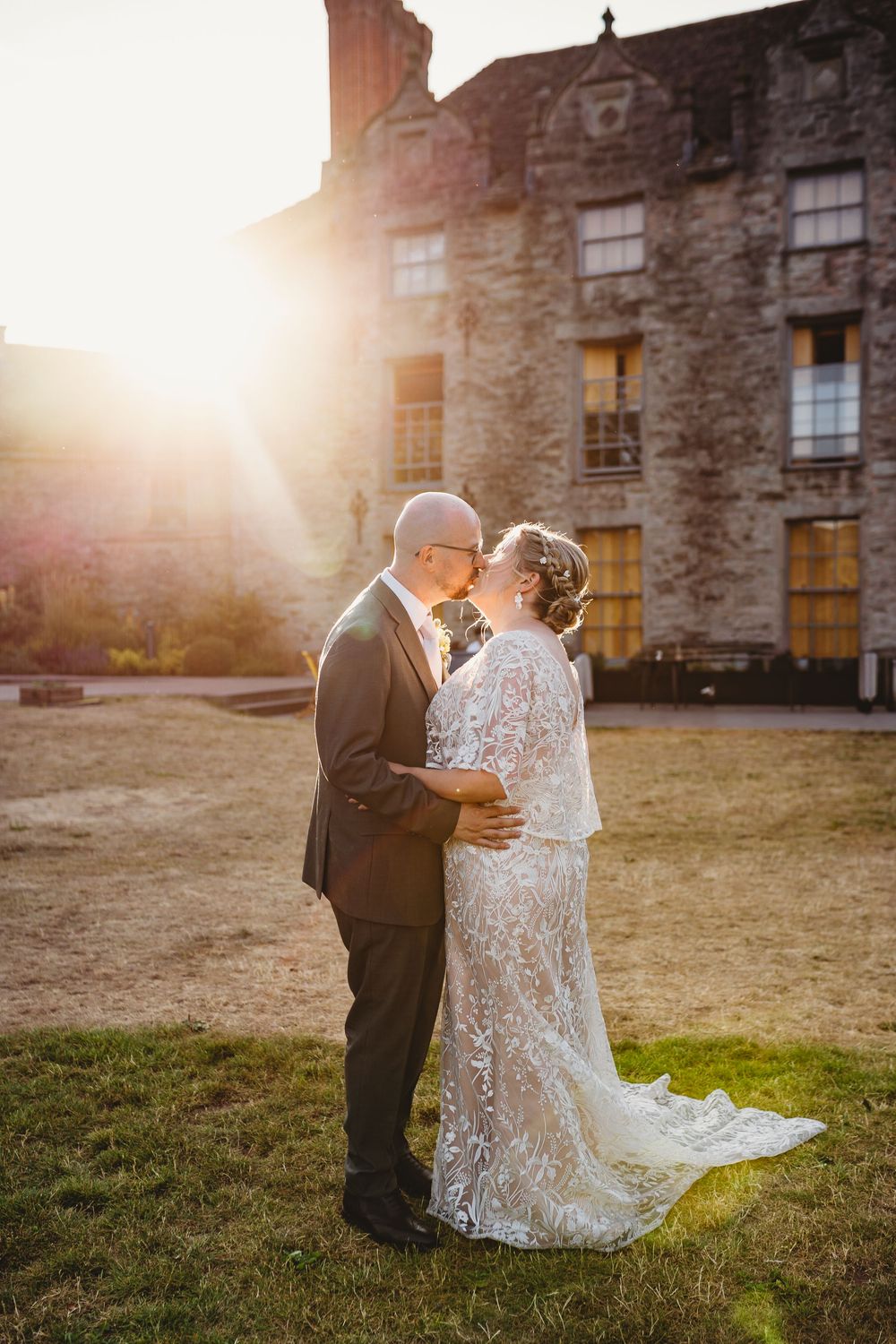 Bride and groom share a romantic sunset kiss outside a historic stone building, bride wearing lace gown with train.