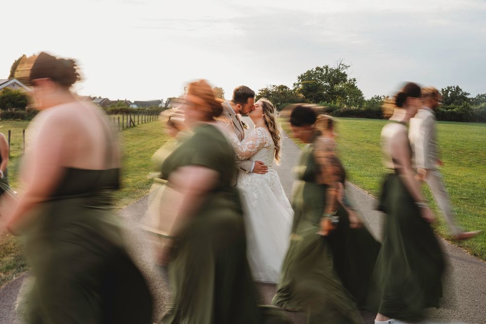 Bride and groom kissing surrounded by blurred bridesmaids in olive green dresses during outdoor wedding celebration.