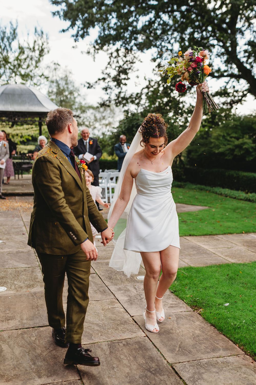 Bride raises colorful bouquet in celebration while holding groom's hand after outdoor wedding ceremony.