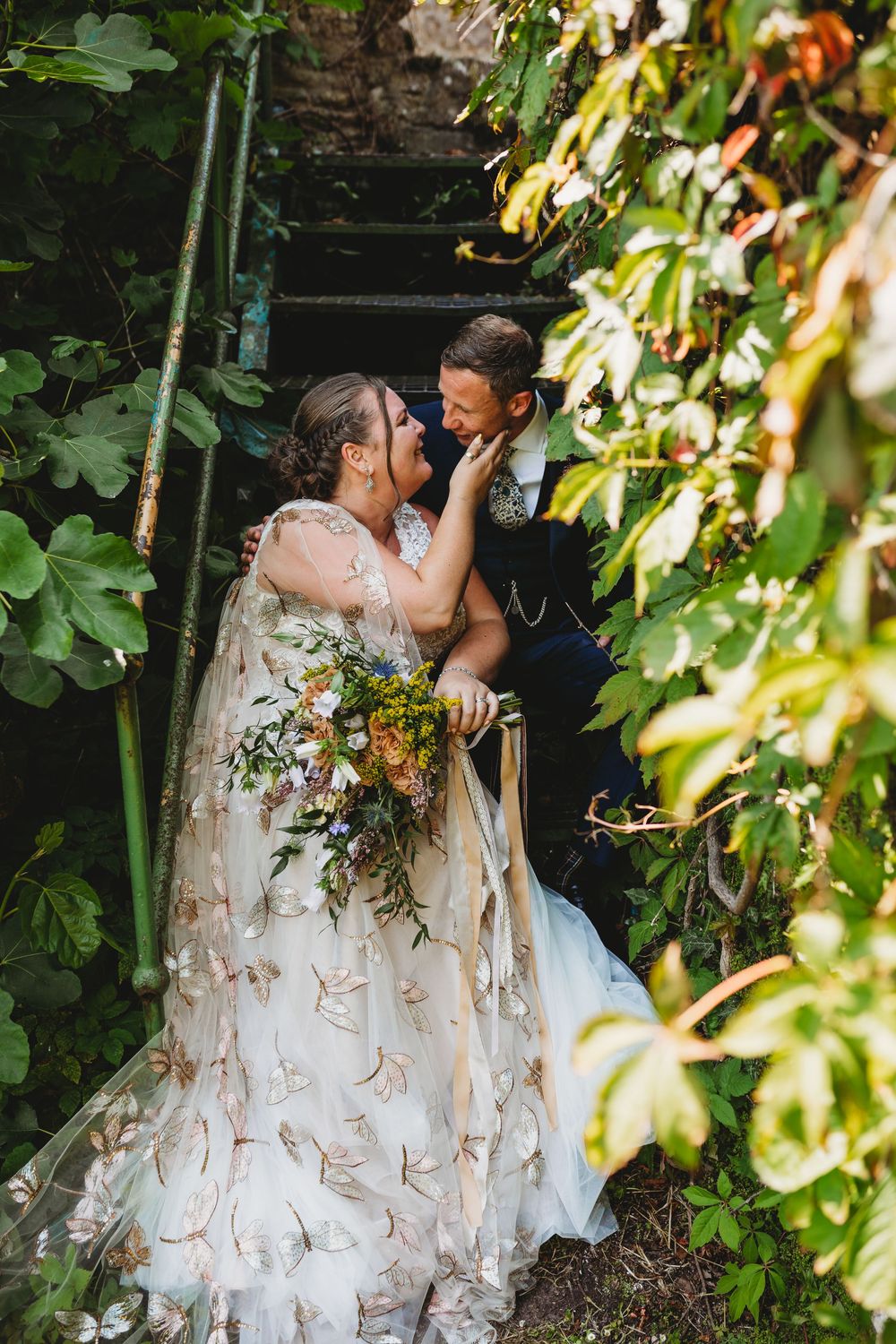 Bride and groom share a romantic moment on ivy-covered stairs, bride holding a wildflower bouquet in a floral gown.
