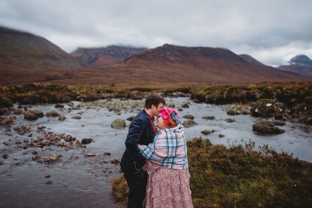 Couple sharing a romantic kiss by a rocky river with misty mountains in the Scottish Highlands backdrop.