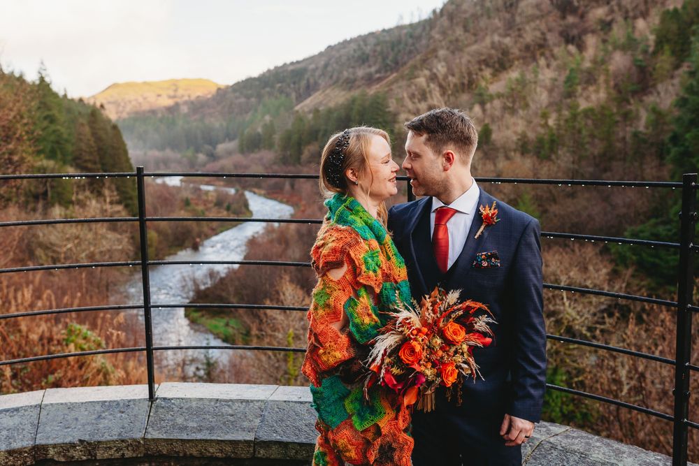 Bride in colorful floral dress and groom in navy suit embrace at scenic overlook with river and autumn hills backdrop.