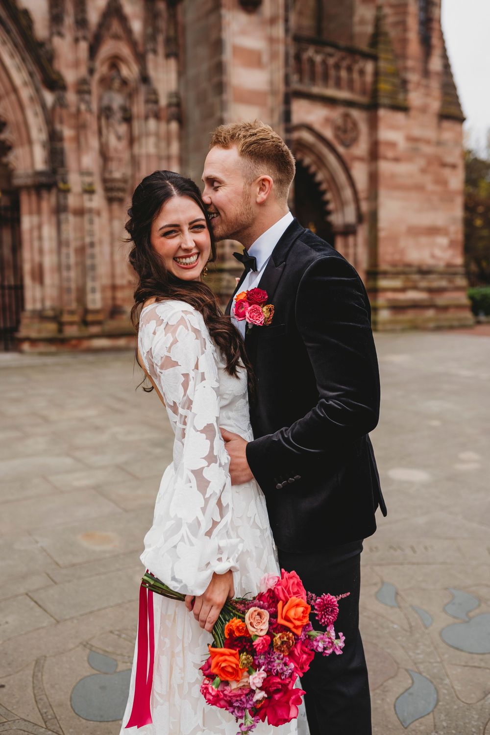 Bride in lace dress holds colorful bouquet while groom in black suit embraces her outside a stone church.