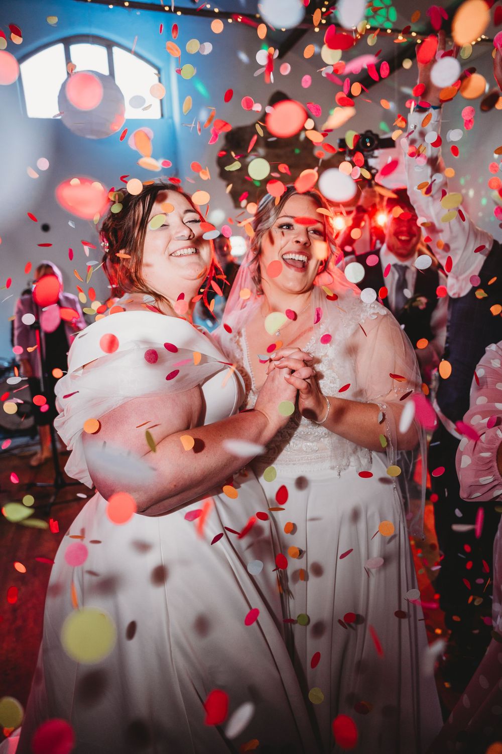 Two brides in white dresses celebrate joyfully surrounded by colorful confetti at their wedding reception.