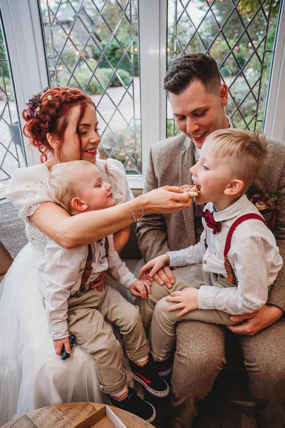 Bride and groom share wedding cake with two young boys in formal attire, seated by a lattice window.