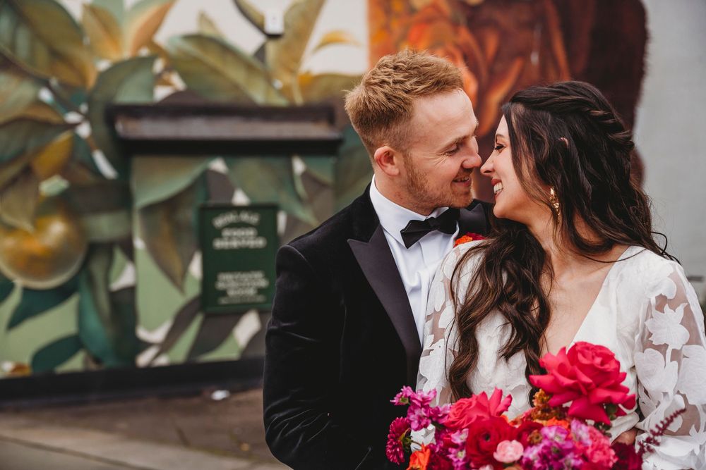 Bride and groom share a romantic moment in front of a colorful mural, bride holding vibrant pink bouquet.