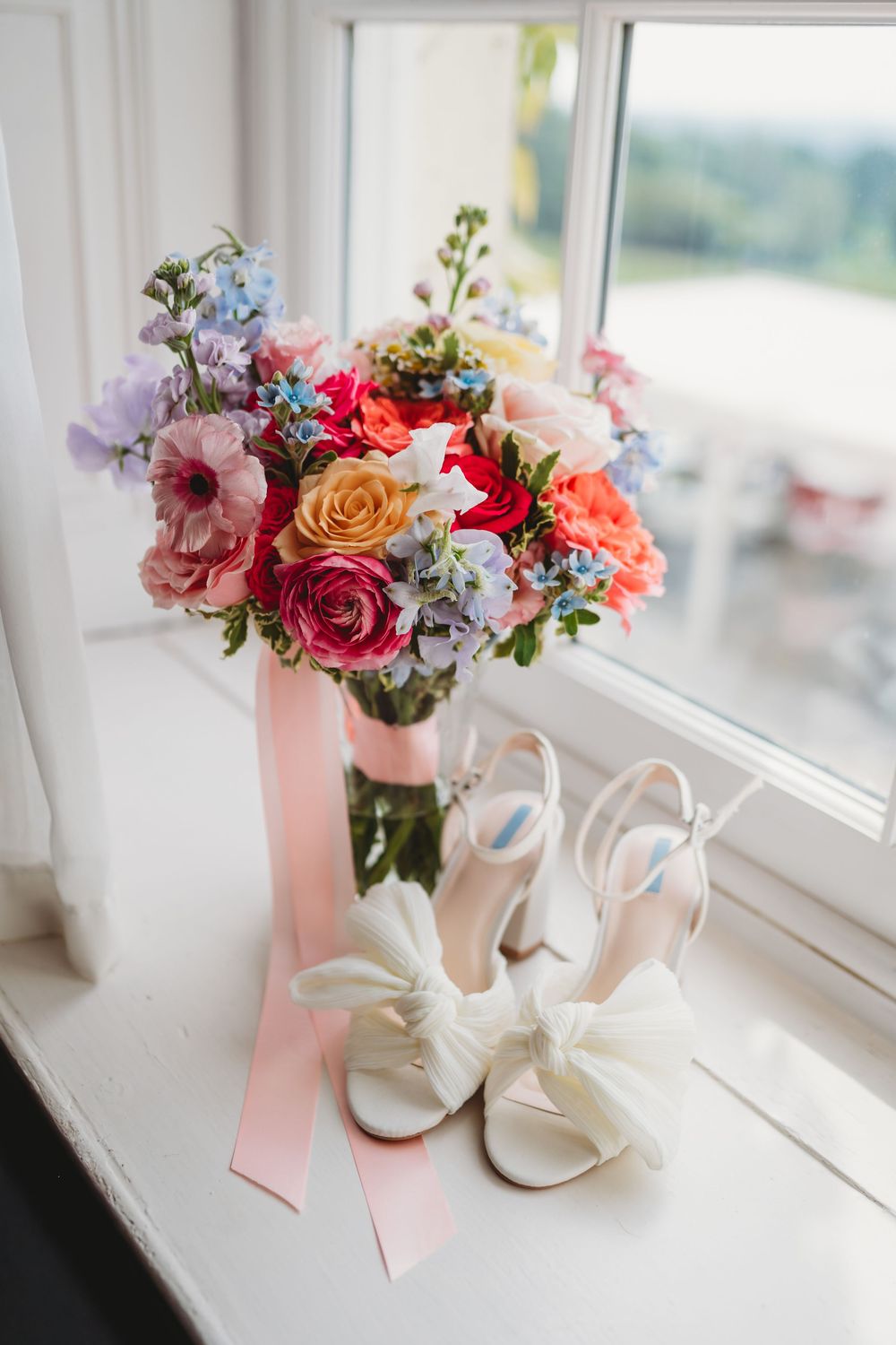 Bridal bouquet with colorful roses and a pink ribbon beside white bow heels on a windowsill.