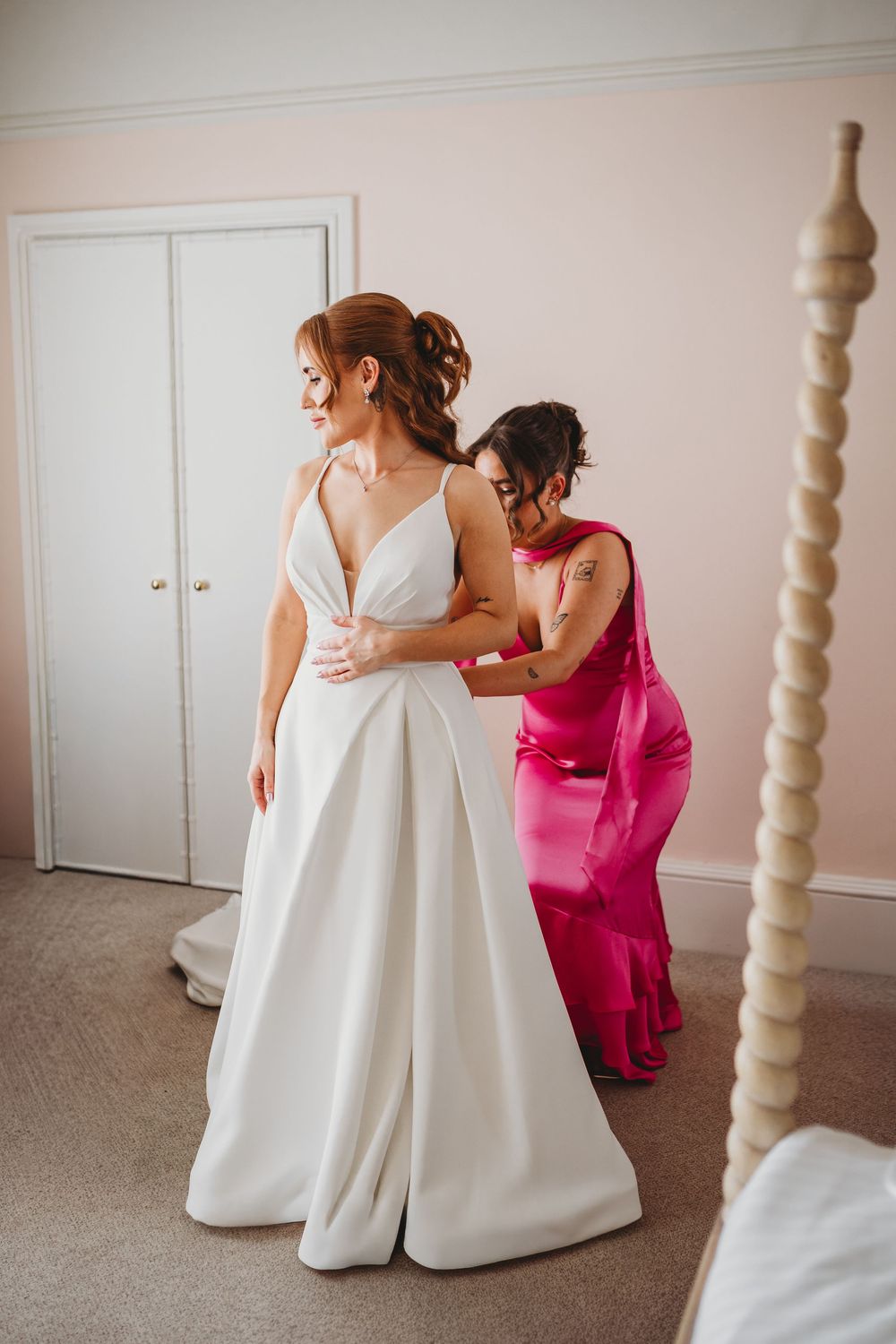 Bridesmaid in pink dress helping bride into elegant white wedding gown in a softly lit bridal suite.