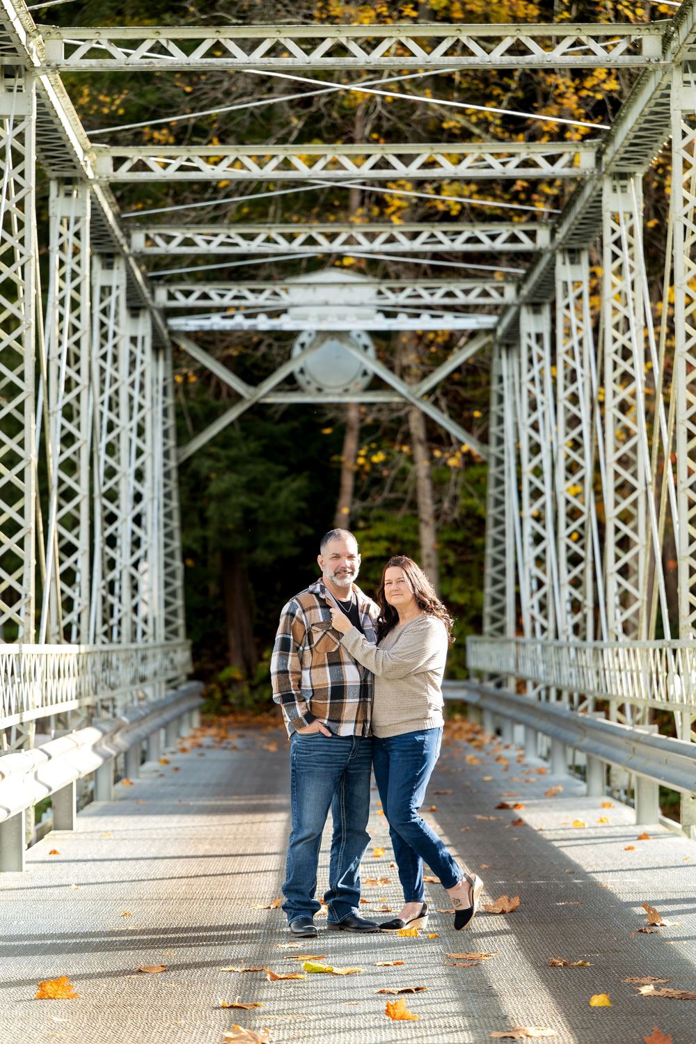 Smiling couple in casual autumn clothing posing together on a steel truss bridge with fallen leaves and fall foliage behind them.