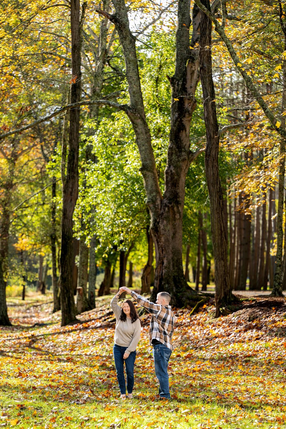 Couple dancing hand in hand on a grassy path surrounded by tall trees and golden autumn leaves in a sunny park.